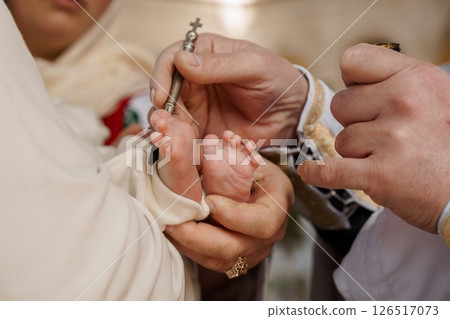 A tender moment during a baby's christening ceremony, showcasing the priest anointing the infant's foot with holy oil, symbolizing spiritual cleansing and entry into the Christian faith A tender moment during a baby's christening ceremony, showcasing the priest anointing the infant's foot with holy oil, symbolizing spiritual cleansing and entry into the Christian faith 126517073