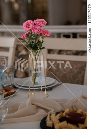 Elegant table setting with pink roses arrangement in a gold vase, white tablecloth, and charcuterie board, creating a romantic and sophisticated atmosphere 126517079