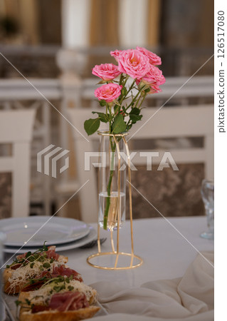 Elegant wedding table setting featuring a delicate pink rose arrangement in a sleek gold vase, complementing a plated appetizer and refined linen decor 126517080