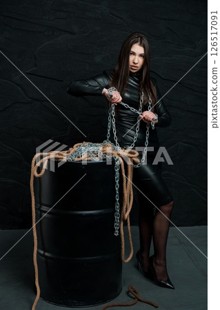 A striking young woman in a black latex catsuit poses confidently with a heavy metal chain and thick rope against a dark textured brick wall, creating a dramatic aesthetic 126517091
