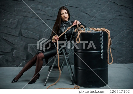 A captivating young woman with long dark hair, dressed in a sleek black leather dress, sits confidently while holding a heavy chain beside a dark barrel A captivating young woman with long dark hair, dressed in a sleek black leather dress, sits confidently while holding a heavy chain beside a dark barrel 126517102