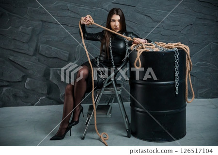 A striking young woman in a black latex catsuit seated on a stool beside a metal barrel, holding ropes and chains in a dimly lit, textured room 126517104