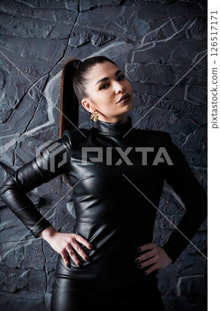 A striking young woman with a determined gaze poses in a sleek black leather dress and matching top against a textured dark stone wall, exuding confidence A striking young woman with a determined gaze poses in a sleek black leather dress and matching top against a textured dark stone wall, exuding confidence 126517171