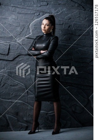 Elegant woman in a tight black leather dress posing confidently with crossed arms against a textured dark stone wall background; sophisticated style Elegant woman in a tight black leather dress posing confidently with crossed arms against a textured dark stone wall background; sophisticated style 126517178