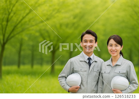 A man and a woman in work clothes are standing smiling and holding helmets in the greenery 126518233