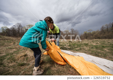 Young adventurers working together to set up a bright orange tent in a beautiful green field under a dramatic sky just before a storm Young adventurers working together to set up a bright orange tent in a beautiful green field under a dramatic sky just before a storm 126519732