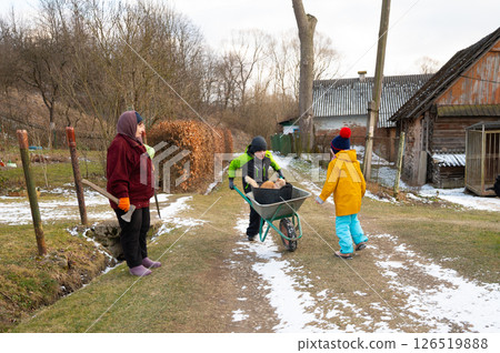 Children playing in the countryside during winter while helping each other with a wheelbarrow on a snowy path surrounded by nature and rustic buildings 126519888