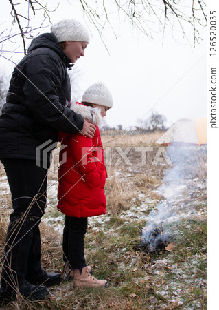 A mother and her daughter are enjoying a peaceful and heartwarming moment together beside a cozy fire while winter camping in a beautiful, snowy field 126520035