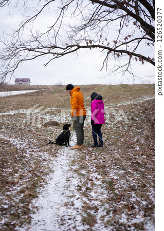 A couple is delighting in a peaceful winter afternoon stroll through the countryside, accompanied by their playful dog in the snow 126520377