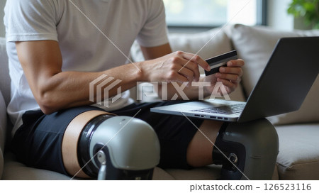 Man with bionic knee prosthesis buying online with credit card and laptop 126523116