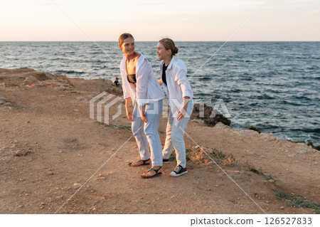 Women Beach Sunset Two women in white clothing stand on a beach cliff at sunset, looking out at the ocean. Women Beach Sunset Two women in white clothing stand on a beach cliff at sunset, looking out at the ocean. 126527833