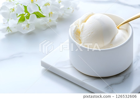 A scoop of pure white vanilla ice cream in an elegant white ceramic bowl on a marble surface. The composition includes delicate white flowers with green leaves in the background, creating a clean. 126532606