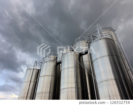 Row of tall silver tanks are lined up in front of a cloudy sky Row of tall silver tanks are lined up in front of a cloudy sky 126532759