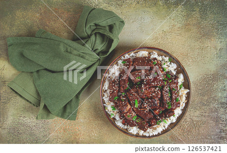 crispy beef, with green onions and sesame seeds, on a rice pad, in a bowl with chopsticks, on a textured surface, no people 126537421