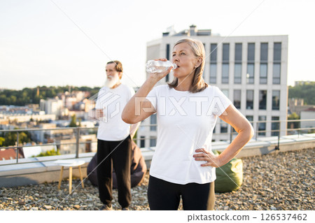 Senior woman drinking water and man standing outside on rooftop, enjoying refreshment. Vibrant composition emphasizing health and fitness. Clear sky and cityscape add energy and inspiration. Senior woman drinking water and man standing outside on rooftop, enjoying refreshment. Vibrant composition emphasizing health and fitness. Clear sky and cityscape add energy and inspiration. 126537462