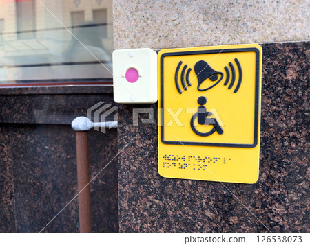 Bright yellow accessibility button for disabled assistance with Braille text and signal icon mounted on granite wall near entrance, promoting inclusive urban infrastructure 126538073