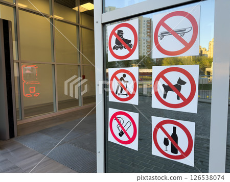 Set of bold prohibition signs on glass door showing banned items like dogs, alcohol, smoking, scooters and skateboards, emphasizing public order and facility regulations 126538074