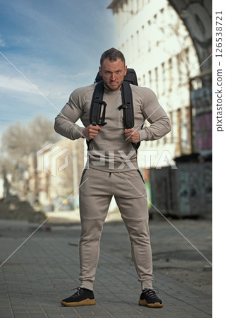 muscular man stands confidently in a casual outfit adjusting his backpack in a city setting under clear skies. He seems ready for an intense exercise session outdoors surrounded by urban elements. 126538721