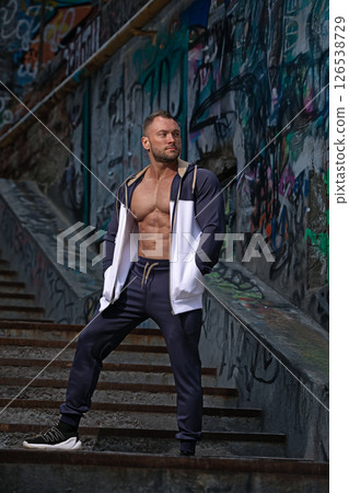 fit man stands on a set of stair steps adorned with vibrant graffiti artwork. He wears a casual outfit and showcases his toned physique while exuding confidence in the daylight. 126538729