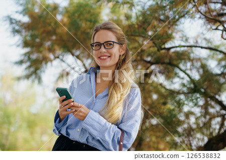 Confident Woman Engaged With Her Smartphone While Outdoors in a Stylish Outfit and Enjoying Life Confident Woman Engaged With Her Smartphone While Outdoors in a Stylish Outfit and Enjoying Life 126538832