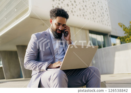 A professional man is utilizing a laptop while on the phone in a modern outdoor environment A professional man is utilizing a laptop while on the phone in a modern outdoor environment 126538842