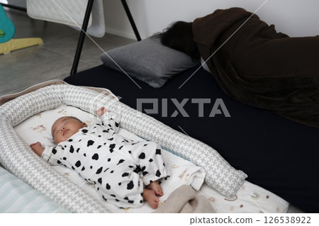 一個正在睡覺的三個月大的嬰兒。一位母親因為養育孩子而精疲力竭,正在後面的蒲團上睡覺。 一個正在睡覺的三個月大的嬰兒。一位母親因為養育孩子而精疲力竭,正在後面的蒲團上睡覺。 126538922