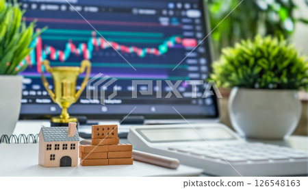 house and bricks on desk with financial chart on screen in background, symbolizing real estate investment and market trends 126548163