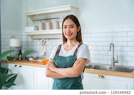 Happy young asian woman wearing apron and standing in kitchen room Happy young asian woman wearing apron and standing in kitchen room 126548982