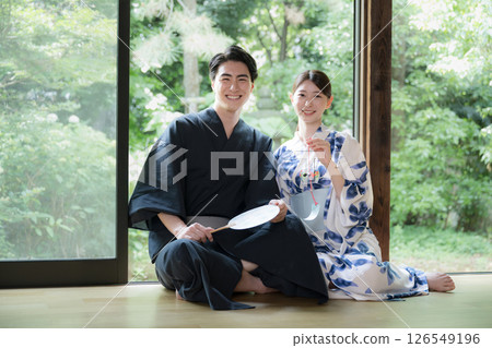 A man and a beautiful woman in yukata relaxing on a veranda surrounded by beautiful greenery. Image of a hot spring after a trip or a stay at a ryokan. Looking at the camera. 126549196
