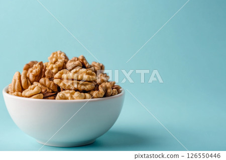 A white bowl filled with walnuts sits on a light blue background. A white bowl filled with walnuts sits on a light blue background. 126550446
