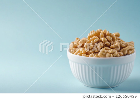 A white bowl filled with walnuts sits on a light blue background. A white bowl filled with walnuts sits on a light blue background. 126550459