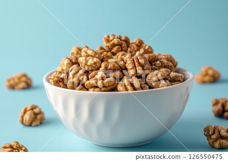 A white bowl filled with walnuts sits on a light blue background. A white bowl filled with walnuts sits on a light blue background. 126550475