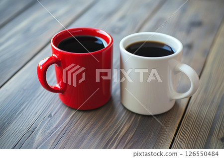 Two ceramic mugs, one red and one white, sit side by side on a wooden table, with a softly blurred background featuring brick walls and greenery. 126550484