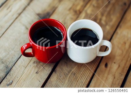 Two ceramic mugs, one red and one white, sit side by side on a wooden table, with a softly blurred background featuring brick walls and greenery. 126550491
