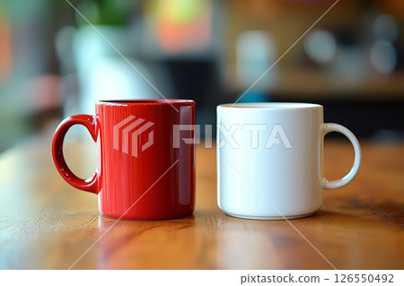 Two ceramic mugs, one red and one white, sit side by side on a wooden table, with a softly blurred background featuring brick walls and greenery. 126550492