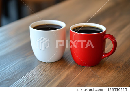 Two ceramic mugs, one red and one white, sit side by side on a wooden table, with a softly blurred background featuring brick walls and greenery. 126550515