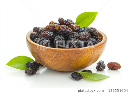 A wooden bowl filled with glossy, dark dates is surrounded by green leaves, with a few dates scattered outside the bowl. 126551604