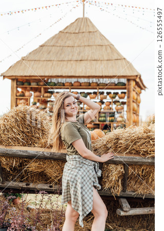 young woman stands at pumpkin festival, holding large orange and green pumpkin. She is surrounded by rows of pumpkins and a wooden display. The autumn farm setting captures the harvest season vibe. young woman stands at pumpkin festival, holding large orange and green pumpkin. She is surrounded by rows of pumpkins and a wooden display. The autumn farm setting captures the harvest season vibe. 126555497