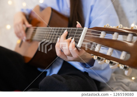 Young woman playing classical guitar during a cozy indoor setting with fairy lights in the background. Fingers with long nails and manicure, strings, neck and head of the guitar close up. Fingerstyle Young woman playing classical guitar during a cozy indoor setting with fairy lights in the background. Fingers with long nails and manicure, strings, neck and head of the guitar close up. Fingerstyle 126559080