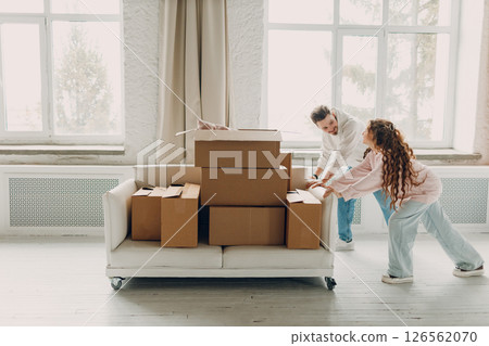 Happy young family couple man and woman push sofa with cardboard boxes in new home. Moving delivery concept. 126562070