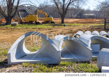 Concrete pipes are in foreground as heavy equipment works in construction zone under works day Concrete pipes are in foreground as heavy equipment works in construction zone under works day 126562101