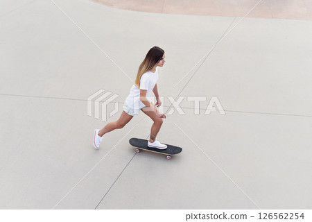 Skateboarder girl in a white t-shirt and denim shorts rides a skateboard in modern urban skatepark. Skateboarder girl in a white t-shirt and denim shorts rides a skateboard in modern urban skatepark. 126562254