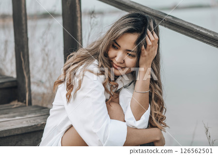 Asian young woman with wavy hair on wooden stairs in white shirt, with hand in hair, peaceful emotional moment outdoors near water, mindfulness, feminine wellness, emotional balance 126562501