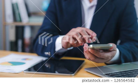 Businessman using smartphone for digital communication or financial analysis at office desk, representing mobile productivity, Technology in business, and online data management. 126564582