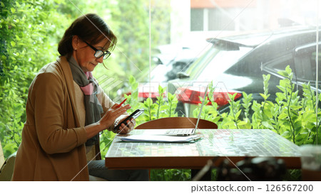 Senior and Technology. Middle aged or senior woman enjoys remote work and online communication technology using various digital devices laptop tablet or smartphone in greeny filled cafe natural light Senior and Technology. Middle aged or senior woman enjoys remote work and online communication technology using various digital devices laptop tablet or smartphone in greeny filled cafe natural light 126567200