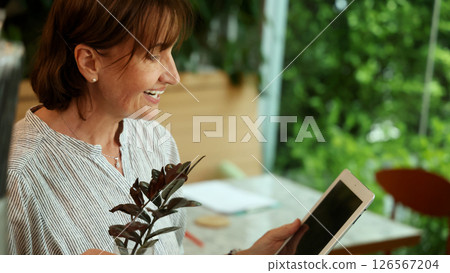 Senior and Technology. Middle aged or senior woman enjoys remote work and online communication technology using various digital devices laptop tablet or smartphone in greeny filled cafe natural light 126567204