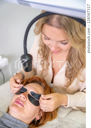 Beautician applying black eye patches before eyelash extension procedure with ultraviolet lamp. Innovative lash extension method in modern beauty salon Beautician applying black eye patches before eyelash extension procedure with ultraviolet lamp. Innovative lash extension method in modern beauty salon 126567317