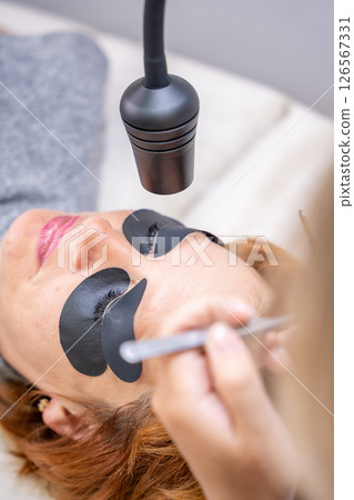 Beautician applying black eye patches before eyelash extension procedure with ultraviolet lamp. Innovative lash extension method in modern beauty salon Beautician applying black eye patches before eyelash extension procedure with ultraviolet lamp. Innovative lash extension method in modern beauty salon 126567331