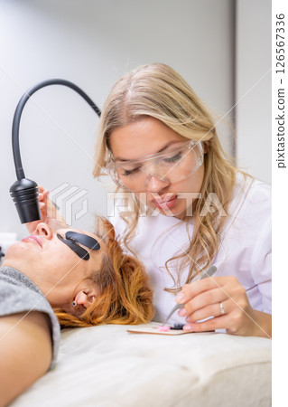 Eyelash extension procedure using ultraviolet lamp in modern beauty salon. Innovative lash enhancement technology with precise application technique. 126567336