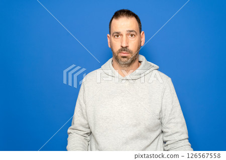 Hispanic man with a beard in his 40s wearing a gray sweatshirt looks straight ahead with a serious and worried expression, isolated on a blue studio background. 126567558